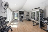 Located at 701 Crest Valley Way in Birmingham, Alabama, this dedicated exercise room delivers a comprehensive fitness environment within a bright, thoughtfully designed space. The room is defined by striking vaulted ceilings with recessed lighting, which create an open, airy atmosphere that enhances the sense of scale. Light hardwood flooring runs throughout, contributing to the clean, modern aesthetic.

Large windows along the walls flood the interior with natural light, offering views of the surrounding greenery and keeping the space feeling fresh and energizing. A white-painted fireplace serves as a focal point on one wall, topped with a wall-mounted television — a practical feature for entertainment during workouts.

The fitness setup is diverse and well-organized. Cardio equipment includes a treadmill and an elliptical/rowing machine, while a multi-station cable weight machine supports strength training. A dedicated dumbbell rack provides a full range of free weights, and a shiplap accent wall serves as a functional ball storage display, holding an assortment of medicine balls and stability balls alongside a stretching mat rack. A large floor fan ensures ventilation and comfort during intense sessions.

Natural light, vaulted ceilings, and a versatile equipment layout make this exercise room a functional and inviting space for a thorough fitness routine.