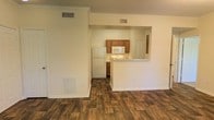 This single-floor space is designed with an open and functional layout that connects the living area to the kitchen through a distinctive pass-through opening in the wall. The living area features rich, wood-look hardwood-style flooring that extends throughout, complemented by crisp white walls and classic crown molding that adds a refined architectural detail. A wall-mounted vent and baseboard heating unit are integrated discreetly into the living space.

The kitchen is visible through the open pass-through, which also doubles as a breakfast bar or counter space, enhancing the sense of connectivity between rooms. It is equipped with warm-toned wooden cabinetry, a white refrigerator, a built-in microwave, and a sink, providing practical storage and workspace. Additional cabinetry is visible below the counter, maximizing storage capacity. A door off the kitchen leads to a separate area of the unit.

Ceiling vents are positioned throughout, ensuring efficient climate control. The overall ambiance is clean, neutral, and move-in ready, with a simple yet functional design that suits a variety of lifestyles.