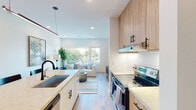 Located at 321 SE Second St in Bentonville, Arkansas, this single-floor unit offers a thoughtfully designed open-concept layout with contemporary finishes throughout. The kitchen serves as a central focal point, featuring warm wood cabinetry that extends to the ceiling, light quartz countertops, a large undermount sink with a matte black faucet, stainless steel appliances, and a white subway tile backsplash. A peninsula with seating and a sleek linear pendant light overhead creates a natural gathering space. A built-in desk nook integrates a convenient workspace seamlessly into the kitchen area. The living area flows directly from the kitchen, bathed in natural light from large windows overlooking tree-lined outdoor views. Light hardwood flooring runs throughout, unifying the space. The bedroom is a calm retreat with natural light from a sizable window dressed in deep navy curtains, a ceiling fan, and a wall-mounted television. The bathroom maintains the clean aesthetic with large-format white tile in the walk-in glass shower, matte black hardware, and light wood flooring. Clean white walls, recessed lighting, and consistent matte black hardware accents lend a cohesive, modern character to the entire space.
