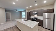 This single-floor residence offers a thoughtfully designed open-concept layout that seamlessly connects the kitchen and living areas. The kitchen serves as the centerpiece of the space, anchored by a generously sized island topped with light granite countertops featuring subtle veining, an integrated sink, and a contemporary gooseneck faucet. Dark espresso-toned cabinetry lines the kitchen walls, providing ample storage, while a white subway tile backsplash adds a clean, classic contrast. A full suite of stainless steel appliances — including a refrigerator, electric range, and microwave — completes the functional cooking space. Recessed lighting and wood-look vinyl flooring extend throughout the kitchen and main living area, which benefits from natural light through a window with blinds and is outfitted with a ceiling fan. The bedroom offers plush carpeting, a ceiling light fixture, and a window that allows additional natural light to flow in. The bathroom features a full soaking tub with a tiled surround, brushed nickel fixtures, wood-look flooring, and clean neutral finishes throughout. The overall aesthetic is modern and cohesive, with a neutral color palette enhancing the sense of openness.