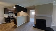 This single-floor space is designed with an open-concept layout that connects the kitchen and living area into one cohesive, functional environment. The kitchen is outfitted with dark espresso cabinetry, stainless steel appliances including a refrigerator, microwave, and range, and a peninsula counter with a sink that serves as a natural divider between the kitchen and the carpeted living area. The transition between the hardwood-style flooring in the kitchen and the carpeting in the living space adds visual contrast while defining each zone. A textured popcorn ceiling runs throughout, complemented by crown molding details that add a subtle decorative touch. The living area features a fireplace with a clean white mantel and dark surround, serving as a prominent focal point. A doorway at the far end of the living space leads to an additional room, where a window allows natural light to filter in, brightening the overall atmosphere. Neutral wall tones throughout create a versatile backdrop, making the space adaptable to a variety of interior styles. The layout is practical and well-organized, offering clear sightlines across the main living areas.