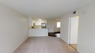 This single-floor space showcases an open-concept design that connects the living area and kitchen in a cohesive, functional layout. The living area is finished with neutral-toned carpeting, creating a soft and understated foundation that complements the overall light color palette of cream and white throughout.

The kitchen is positioned along a peninsula-style counter that separates it from the main living space while maintaining visual openness. It features white cabinetry, a stainless steel refrigerator, and a built-in sink along the countertop, providing practical workspace and storage. Under-cabinet lighting adds a functional touch to the cooking area.

Beyond the kitchen, a secondary room is visible, distinguished by its dark hardwood-style flooring, offering a contrast in texture and finish from the carpeted living area. A window in this room allows natural light to filter through, brightening the interior. The space benefits from consistent white walls and ceilings throughout, enhancing the sense of brightness and openness. A doorway to the right of the living area suggests additional rooms, contributing to the overall functional flow of the layout.
