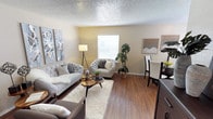 This single-floor space showcases a thoughtfully arranged open-concept layout that combines a living area and dining space. The flooring throughout is warm-toned hardwood, lending a cohesive and inviting feel to the entire space. A textured popcorn ceiling adds a classic architectural detail overhead.

The living area is generously proportioned, accommodating a tufted sofa, an accent armchair, and a mid-century modern coffee table set atop a plush white area rug. Decorative wall art flanks both sides of the room, adding visual interest and personality. A tall floor lamp provides supplemental lighting alongside a large window dressed with white blinds, which allows natural light to filter into the space.

Greenery in the form of a tall potted plant adds a natural, organic element near the window. The dining area is positioned adjacent to the living space and features bar-height seating alongside a dark credenza, creating a functional yet stylish transition between the two zones. The neutral wall tones throughout contribute to an airy, calm ambiance that makes the space feel open and welcoming.
