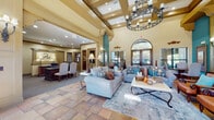 Located at 215 N Loop 1604 E Access Rd in San Antonio, Texas, this single-story recreation center offers a comprehensive suite of community amenities wrapped in a warm Southwestern-inspired design. The grand main lobby immediately impresses with soaring coffered ceilings, a wrought-iron chandelier, and large arched windows that flood the space with natural light. Comfortable seating arrangements with plush sofas and accent pillows create an inviting lounge atmosphere, while a dedicated seating area with a round wooden table and upholstered chairs provides a more intimate setting. Rich terracotta tile flooring flows throughout the main areas, complemented by teal accent walls and warm beige tones. A fully equipped community kitchen features bold teal cabinetry, granite countertops, stainless steel appliances, and hardwood flooring — ideal for gatherings and events. The fitness center is outfitted with a broad selection of cardio and strength-training equipment beneath a tray ceiling with teal accents and floor-to-ceiling mirrors. A dedicated business suite with built-in cabinetry and a workstation offers a quiet workspace, while a tiled corridor lined with artwork connects the facility's various wings. A clean, well-maintained laundry room with multiple washing machines rounds out the extensive amenities of this community facility.