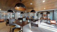 This single-level co-working and business lounge is designed with a sophisticated blend of modern and industrial aesthetics. The ceiling is clad in rich, warm wood planks that add texture and visual depth throughout the space, complemented by oversized black dome pendant lights with copper interiors that provide both ambient and task lighting.

The flooring features a herringbone-patterned carpet tile in neutral tones, lending a refined, contemporary feel underfoot. Structural columns wrapped in light-toned brick add an industrial character while anchoring the open layout. The space is organized into distinct functional zones: a dedicated workstation area along the perimeter wall includes built-in desks equipped with desktop computers and task seating, ideal for focused individual work. Multiple bar-height communal tables with metal stools are centrally positioned, encouraging collaborative or casual work sessions.

A fully enclosed glass-walled conference room provides a private meeting space, visible through sleek black-framed floor-to-ceiling glass panels and outfitted with a wall-mounted display screen. Floor-to-ceiling windows along one exterior wall flood the space with natural light and frame views of lush landscaping and greenery outside. Accent seating areas with upholstered chairs are tucked near the windows, offering relaxed retreat spots within the broader workspace environment.