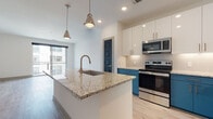 This thoughtfully designed single-floor residence offers a clean, contemporary aesthetic throughout. The open-concept layout connects the kitchen and living area seamlessly, creating an inviting and functional space. The kitchen is a standout, featuring a large central island with granite countertops and an undermount sink with a brushed gold faucet, illuminated by sleek pendant lights. Two-tone cabinetry — bold teal lowers paired with crisp white uppers — adds a striking design element, while a subway tile backsplash and a full suite of stainless steel appliances complete the look. Light hardwood flooring flows throughout the main living areas, enhancing the sense of openness and warmth. The spacious living area is bathed in natural light through large sliding glass doors that open to a private balcony with exterior views. The bedroom features plush carpeting, a ceiling fan, and dark-paneled interior doors that lend a bold contrast to the neutral walls. The bathroom is finished with a tiled soaking tub and shower combo, brushed nickel fixtures, a teal vanity with granite countertop, and light wood-style flooring, creating a clean and cohesive look. Dark interior doors and recessed lighting are consistent design details found throughout the residence.