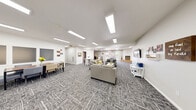 Located in St. George, Utah, this single-floor community space offers a generously sized, open-plan layout designed to accommodate a variety of group activities and social gatherings. The interior is finished with a neutral color palette of crisp white walls and patterned gray carpet throughout the main areas, creating a clean and welcoming atmosphere.

The space is divided into several functional zones. A comfortable lounge area features a gray sofa set, wall-mounted television, and decorative wall art. Adjacent to it, a dining zone provides seating at multiple tables, suitable for shared meals or group events. A dedicated dining nook near the windowed wall is brightened by natural light from multiple windows fitted with roller shades. A kitchen area with tile flooring, a breakfast bar with bar stools, cabinetry, and appliances adds practical utility to the space.

A white-surround fireplace serves as a focal point along one wall, flanked by windows that overlook an outdoor patio accessible through glass French doors — providing views of the exterior pool area. Overhead, an array of recessed rectangular panel lights ensures bright, even illumination throughout. The space also includes direct access to additional rooms and exterior exits, making it highly functional for community use.
