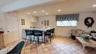This single-story clubhouse offers a comfortable and functional gathering space designed for socializing and entertaining. The main area is anchored by a white L-shaped bar counter equipped with an overhead ventilation hood, making it well-suited for food and beverage service during events. Black wicker bar stools with cushioned seats line the counter, providing ample seating for guests.

The flooring throughout the space consists of warm terracotta square tiles, lending a casual, inviting character to the interior. Recessed lighting installed in the ceiling ensures even, ambient illumination across the room. A large window dressed with black-and-white striped balloon curtains allows natural light to filter in while adding a classic decorative touch.

The neutral, cream-toned walls serve as a clean backdrop, accented by framed artwork arranged in gallery-style groupings on either side of the room. Additional seating, including a patterned chair, is positioned near the window, creating a relaxed lounge area. The overall ambiance is bright, tidy, and functional, making this clubhouse an ideal venue for community gatherings and events.