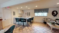 This single-story clubhouse offers a comfortable and functional gathering space designed for socializing and entertaining. The main area is anchored by a white L-shaped bar counter equipped with an overhead ventilation hood, making it well-suited for food and beverage service during events. Black wicker bar stools with cushioned seats line the counter, providing ample seating for guests.

The flooring throughout the space consists of warm terracotta square tiles, lending a casual, inviting character to the interior. Recessed lighting installed in the ceiling ensures even, ambient illumination across the room. A large window dressed with black-and-white striped balloon curtains allows natural light to filter in while adding a classic decorative touch.

The neutral, cream-toned walls serve as a clean backdrop, accented by framed artwork arranged in gallery-style groupings on either side of the room. Additional seating, including a patterned chair, is positioned near the window, creating a relaxed lounge area. The overall ambiance is bright, tidy, and functional, making this clubhouse an ideal venue for community gatherings and events.