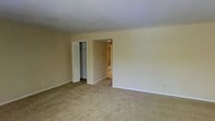 This single-floor space presents a straightforward and functional layout. The primary room shown is generously sized, finished with wall-to-wall beige carpeting that adds a sense of warmth underfoot. Neutral cream-colored walls and a clean white ceiling create a bright, understated backdrop throughout the space.

A built-in closet with bifold doors is positioned along one wall, offering convenient storage directly within the room. The closet interior features a visible hanging rod, providing organized space for clothing or other items.

An open doorway leads to an adjoining area, where a transition to hard-surface flooring is visible, suggesting a distinct functional zone such as a hallway, kitchen, or utility area. The passageway is well-lit, hinting at additional rooms beyond.

The overall ambiance of the space is clean and neutral, lending itself well to personalization. The simple architectural details, including white baseboards and standard ceiling height, contribute to a practical and livable environment. The absence of clutter and the use of light, neutral tones throughout give the space an open and airy feel, making it easy to envision a variety of furnishing arrangements.