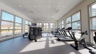 Located at 2155 Morris Basket Blvd in North Charleston, SC, this single-story fitness center offers a spacious, thoughtfully designed workout environment. The facility is enclosed by large, multi-pane windows along multiple walls, flooding the interior with natural light and providing views of the surrounding landscaping and community grounds, including glimpses of a pool area. Ceiling fans and recessed lighting complement the bright, open atmosphere throughout.

The main workout floor features durable speckled rubber flooring suited for high-traffic exercise use. Cardio equipment includes a row of treadmills and stationary bikes positioned to take advantage of the exterior views. A dedicated strength training section houses a multi-station cable and resistance machine, along with additional selectorized equipment for targeted muscle training. A free weight area offers a full rack of dumbbells in varying weights, accompanied by adjustable benches and a large wall-mounted mirror that enhances the sense of space. A wall-mounted television provides entertainment during workouts. A hydration station is conveniently accessible near the entrance, and ceiling fans ensure comfortable airflow throughout the facility.