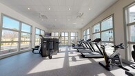 Located at 2155 Morris Basket Blvd in North Charleston, SC, this single-story fitness center offers a spacious, thoughtfully designed workout environment. The facility is enclosed by large, multi-pane windows along multiple walls, flooding the interior with natural light and providing views of the surrounding landscaping and community grounds, including glimpses of a pool area. Ceiling fans and recessed lighting complement the bright, open atmosphere throughout.

The main workout floor features durable speckled rubber flooring suited for high-traffic exercise use. Cardio equipment includes a row of treadmills and stationary bikes positioned to take advantage of the exterior views. A dedicated strength training section houses a multi-station cable and resistance machine, along with additional selectorized equipment for targeted muscle training. A free weight area offers a full rack of dumbbells in varying weights, accompanied by adjustable benches and a large wall-mounted mirror that enhances the sense of space. A wall-mounted television provides entertainment during workouts. A hydration station is conveniently accessible near the entrance, and ceiling fans ensure comfortable airflow throughout the facility.