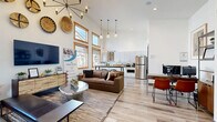 Located at 515 Aurora in Bend, Oregon, this single-floor community space offers a thoughtfully designed open-plan layout that seamlessly integrates a living area and kitchen. The interior immediately impresses with dramatically soaring ceilings that amplify the sense of space and openness. A floor-to-ceiling stacked stone fireplace serves as a striking focal point in the living area, anchoring the room with a warm, rustic elegance. Large wood-trimmed windows flood the space with abundant natural light, offering views of the surrounding neighborhood. Light-toned hardwood flooring runs throughout, complementing the crisp white walls and warm wood accents. The living area is appointed with a built-in window bench, decorative woven wall art, a statement sputnik chandelier, and globe pendant lights, creating an inviting and contemporary ambiance. A dedicated workspace with an adjacent private office provides functional versatility. The kitchen features sleek white cabinetry, granite countertops with stainless steel appliances, and black hardware accents, combining practicality with modern style. Every detail of this space reflects a refined, community-oriented design sensibility, making it an ideal gathering and collaboration hub in the heart of Bend.