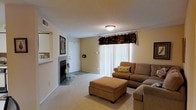 This single-floor space centers around a comfortable living room that balances functionality with a warm, inviting atmosphere. The room is finished with wall-to-wall neutral carpeting that contributes to a soft, cohesive aesthetic throughout. A tile-surround fireplace is positioned along one wall, accompanied by built-in shelving niches that provide display or storage opportunities and serve as a notable architectural detail.

Natural light enters the space through a large sliding glass door dressed with vertical blinds and a decorative dark floral valance, lending a traditional touch to the room's overall character. The entry door is conveniently located nearby, offering a practical flow between the entrance and main living area. A flush-mount ceiling light provides overhead illumination, complementing the light that filters in from the window treatment.

The living area comfortably accommodates a sectional sofa and a large upholstered ottoman, illustrating the generous scale of the room. Neutral beige walls keep the space feeling open and adaptable. Ceiling-mounted ventilation is also present, ensuring year-round comfort. Overall, the space offers a straightforward, well-maintained layout suited for everyday living.