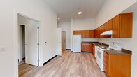 This single-floor unit offers a thoughtfully organized layout across four distinct areas, making efficient use of its compact footprint. The open kitchen is a functional highlight, featuring warm medium-toned wood cabinetry that extends to the ceiling for ample storage, dark countertops, a white appliance suite including an electric range and a full-size refrigerator, and a stainless steel sink. The tall ceilings in the kitchen and living area lend a sense of openness and airiness that extends throughout the space. Light wood-tone laminate flooring runs consistently across the main living and sleeping areas, complemented by clean white walls and bold dark baseboards that provide a sharp, modern contrast. The bedroom is generously appointed with two sliding-door closets — one standard and one fitted with multi-tier wire shelving — offering excellent storage versatility. The bathroom features a vanity with a wood cabinet base and dark countertop, a large mirror, and a full tub/shower combination. The tile-style flooring adds a clean, practical finish to the bathroom. Recessed ceiling lighting illuminates the main areas, while the overall aesthetic is modern, minimal, and move-in ready.