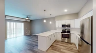 This single-floor residence offers a thoughtfully designed open-concept layout that seamlessly connects the kitchen and living areas. The kitchen is a standout feature, showcasing crisp white shaker cabinetry, a suite of stainless steel appliances including a refrigerator, electric range, microwave, and dishwasher, and a generous peninsula with a bright white countertop that provides additional workspace and casual seating. Elegant pendant lights hang above the peninsula, complementing the recessed lighting throughout. Wood-look plank flooring anchors the kitchen and living space, adding warmth to the contemporary aesthetic. The living area benefits from a ceiling fan and access to a patio or balcony via a sliding glass door, inviting natural light into the open space. The bedrooms are finished with plush carpet flooring, neutral taupe walls, ceiling fans, and windows with blinds that provide a comfortable, restful atmosphere. The bathroom features a wide vanity with a white countertop and undermount sink, a large mirror, and tiled accents, offering a clean and functional design. Throughout the residence, neutral tones and modern finishes create a cohesive and inviting ambiance.