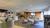 This well-appointed single-floor space offers a thoughtfully designed environment that blends comfort with functionality. The main lounge area features a contemporary layout with plush neutral-toned seating, including accent chairs with ottomans arranged around a round coffee table, creating an inviting space for relaxation or informal meetings. A patterned area rug anchors the seating arrangement, adding visual interest to the tiled flooring beneath.

A warm wood-paneled accent wall serves as a focal point in the room, hosting a large flat-screen display — ideal for presentations or informational content. Adjacent to the lounge, a dedicated collaborative zone offers additional seating with sleek, modern chairs gathered around work-friendly tables, suitable for group discussions or individual use.

The space is illuminated by recessed ceiling lighting set within a clean drop-tile ceiling, providing consistent, comfortable brightness throughout. A glass-partitioned wall offers a seamless visual connection to the adjacent fitness facility, which is visible in the background and appears to be well-equipped with exercise equipment. The open layout and neutral color palette of whites, grays, and warm wood tones contribute to a professional yet welcoming ambiance throughout the entire space.