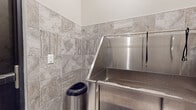 This single-room dog wash facility is thoughtfully designed to provide a clean, functional, and durable space for pet grooming. The room is accessed through a distinctive glass-paneled door etched with paw print graphics and "Dog Wash" signage, making the space easily identifiable. The interior walls are fully clad in large-format marble-look ceramic tiles in warm beige and gray tones with natural veining patterns, lending the space a polished, upscale appearance while remaining highly practical and easy to clean. The same tile treatment extends from floor to ceiling, creating a cohesive and hygienic environment. The centerpiece of the room is a professional-grade, full-size stainless steel dog bathing tub, featuring high surrounding panels to contain splashing, integrated safety leash loops, and a mounted showerhead with an adjustable caddy for grooming products. A stainless steel step or ramp area at the front of the tub provides easy pet access. A sleek stainless steel trash receptacle sits conveniently beside the tub. Electrical outlets are positioned along the tiled walls to accommodate grooming tools such as dryers or clippers. The space is well-lit with overhead lighting, and the compact, efficient layout ensures that everything needed for a thorough wash is within easy reach.
