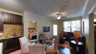 This single-floor residence offers a thoughtfully designed open-concept layout that seamlessly connects the living, dining, and kitchen areas. The living room is warm and inviting, anchored by rich hardwood flooring, a ceiling fan with a light fixture, and crown molding that adds a polished architectural detail throughout the space. A wall-mounted flat-screen television serves as a focal point, while large windows and a glass door leading to a balcony flood the interior with natural light and offer views of the surrounding area. The seating area is generously sized, accommodating a sofa and accent chairs styled in a vibrant orange and white palette that lends the space a lively, contemporary character. A glass-top dining table with patterned chairs provides a defined dining area that flows naturally from the kitchen. The kitchen features dark espresso cabinetry, a tile backsplash in warm earth tones, a stainless steel sink, and ample counter and storage space. The bedroom, visible through a framed doorway, is a comfortable retreat with a dark wood bed frame and coordinating bedding in orange and white tones, continuing the cohesive design aesthetic found throughout the home. Crown molding and neutral wall tones unify each area of this well-maintained space.