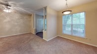 This single-floor residence offers a thoughtfully arranged layout that balances open communal areas with private retreats. The living and dining areas are connected by an open flow, distinguished by decorative white columns that add a subtle architectural detail. The living room features plush carpeting, a ceiling fan with integrated lighting, and windows that invite natural light and outdoor views. A pendant light fixture marks the dining area, which also benefits from generous window coverage. A glass-paneled exterior door provides additional natural light and outdoor access.

The galley-style kitchen is efficiently organized, featuring white upper and lower cabinetry, white countertops, a full-size refrigerator, an electric range, and dark hardwood-style flooring that contrasts cleanly with the bright cabinetry. A doorway at the kitchen's end leads to a utility or storage area with visible shelving and an electrical panel.

The bedroom is carpeted and equipped with a ceiling fan, offering a comfortable and quiet atmosphere. It is served by a spacious walk-in closet outfitted with multiple wire shelving units and hanging rods for ample storage. The bathroom features dark hardwood-style flooring, a wide vanity with white cabinetry, a large mirror, a lighted fixture, and a window for natural light.