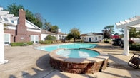Located at 2422 Banks Edge Way in Reynoldsburg, Ohio, this single-floor community amenity space is centered around a generously sized outdoor swimming pool that serves as its defining feature. The pool area is complemented by a raised circular spa with classic brick surround detailing, offering a relaxing retreat alongside the main pool. The surrounding concrete patio provides ample space for lounge chairs, ideal for enjoying warm-weather days under open skies. The complex is framed by charming brick and white-sided structures, including what appears to be a clubhouse or community building with arched windows and a distinctive cupola, lending the space a traditional, well-maintained aesthetic. Auxiliary buildings, including what appears to be a pool house with restroom or storage facilities, are conveniently positioned nearby. Mature trees and established landscaping border the fenced perimeter, creating a sense of privacy and natural greenery. The overall ambiance is welcoming and resort-like, combining classic architectural character with practical outdoor amenities designed for community enjoyment and relaxation.