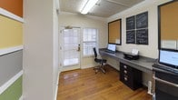 This single-floor business center is a well-appointed and functional workspace designed to meet everyday professional needs. The room features warm hardwood flooring throughout, lending a polished and inviting atmosphere to the otherwise compact, efficient layout. A built-in wall-length desk in a dark charcoal finish provides ample workspace, accommodating a desktop computer, keyboard, and printer — making it a fully equipped station for productivity. Drawers integrated beneath the desk offer convenient storage solutions. A sleek ergonomic office chair completes the workstation setup. Natural light filters into the space through a window fitted with white blinds, while overhead fluorescent lighting ensures consistent illumination. Crown molding along the ceiling adds a refined architectural touch, and a textured ceiling contributes subtle visual interest. One of the standout features of the space is a boldly striped accent wall displaying a curated selection of paint color swatches — including shades like orange, yellow, gray, green, and blue — serving as an interactive design showcase. A large cork bulletin board and two framed chalkboard panels mounted on the wall provide space for notes and announcements. Framed artwork on neutral-toned walls adds a welcoming, professional character to the overall environment.