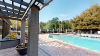 This outdoor pool area offers a well-appointed, resort-style retreat designed for both relaxation and socializing. At the heart of the space is a generously sized swimming pool with clear, blue water, surrounded by a classic herringbone-patterned brick deck that extends throughout the entire area, providing ample room for lounging and movement.

A prominent pergola structure with textured stone columns runs along one side of the deck, offering partial shade and defining a covered patio zone adjacent to the main building. This covered area is furnished with bistro-style seating and accent tables, creating a comfortable spot to unwind. Raised planter beds and large decorative pots filled with colorful flowering plants and tropical foliage add vibrant touches of greenery throughout the space.

The pool is enclosed by a wrought iron fence, ensuring privacy and security while maintaining open sightlines to the surrounding landscape of mature trees, including tall pines and palms, which create a natural and serene backdrop. Additional seating clusters, lounge chairs, and shaded umbrella areas are distributed around the pool perimeter, providing versatile options for outdoor enjoyment. Overhead lighting fixtures ensure the area remains functional and inviting into the evening hours. The overall ambiance is bright, open, and welcoming, with abundant natural light enhancing the lush, park-like setting.