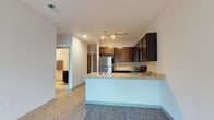 This single-story unit offers a thoughtfully designed open-concept layout that seamlessly connects the kitchen and living areas. The kitchen serves as a central focal point, featuring rich dark-toned cabinetry that contrasts elegantly with light granite countertops. A peninsula with an integrated sink extends into the living space, providing both additional prep space and a natural division between the kitchen and the rest of the unit. Stainless steel appliances, including a refrigerator, range, and dishwasher, complement the modern aesthetic, while a tile backsplash adds a refined finishing touch. Track lighting above the kitchen illuminates the space effectively. Light wood-look luxury vinyl flooring runs throughout, adding warmth and continuity across the open floor plan. Recessed lighting in the main area contributes to a bright, clean ambiance. A hallway visible from the kitchen leads to the bedroom, which appears spacious and includes a bathroom featuring cabinetry consistent with the kitchen's design. The unit balances contemporary style with functional design, making efficient use of its well-proportioned single-floor footprint.