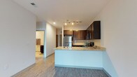 This single-story unit offers a thoughtfully designed open-concept layout that seamlessly connects the kitchen and living areas. The kitchen serves as a central focal point, featuring rich dark-toned cabinetry that contrasts elegantly with light granite countertops. A peninsula with an integrated sink extends into the living space, providing both additional prep space and a natural division between the kitchen and the rest of the unit. Stainless steel appliances, including a refrigerator, range, and dishwasher, complement the modern aesthetic, while a tile backsplash adds a refined finishing touch. Track lighting above the kitchen illuminates the space effectively. Light wood-look luxury vinyl flooring runs throughout, adding warmth and continuity across the open floor plan. Recessed lighting in the main area contributes to a bright, clean ambiance. A hallway visible from the kitchen leads to the bedroom, which appears spacious and includes a bathroom featuring cabinetry consistent with the kitchen's design. The unit balances contemporary style with functional design, making efficient use of its well-proportioned single-floor footprint.