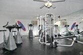 This fitness center is designed to support a comprehensive workout experience within a single-level layout. The room is anchored by bold orange accent walls that create an energizing atmosphere, complemented by a neutral-toned tile floor and a textured ceiling fitted with two ceiling fans that provide continuous air circulation throughout the space.

The facility is stocked with a variety of exercise equipment to accommodate both cardio and strength training needs. Cardio machines include treadmills, ellipticals, and a stationary bike, while a multi-station cable and weight machine supports full-body strength conditioning. The equipment is arranged to allow comfortable movement between stations.

A large mirrored wall spans one side of the room, visually expanding the space and allowing users to monitor their form during workouts. A window on the far wall introduces natural light, brightening the interior and providing a view to the outside. The overall layout is functional and open, making efficient use of the available floor space while maintaining a motivating and practical environment for fitness activity.