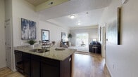 This single-floor residence offers a thoughtfully designed open-concept layout that seamlessly connects the kitchen, dining, and living areas. The kitchen is appointed with dark espresso cabinetry, granite countertops, black appliances, and a peninsula with bar seating — ideal for casual dining. Hardwood-style flooring flows throughout the main living areas, adding warmth and continuity to the space. The living room is generously sized, bathed in natural light through a large sliding glass door that opens to an outdoor balcony, offering views of the surrounding area. Neutral wall tones and a textured ceiling complement the open, airy atmosphere. The bedroom features plush carpeting, a large window that invites natural light, and ample space for a full furniture arrangement. The bathroom is well-equipped with a granite-topped dark vanity, a large mirror with bright vanity lighting, a combined tub and shower, and tile flooring. A dedicated in-unit laundry area houses a full-sized washer and dryer with overhead shelving for added storage. Throughout the residence, the design balances practical functionality with a clean, contemporary aesthetic.