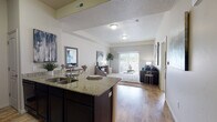 This single-floor residence offers a thoughtfully designed open-concept layout that seamlessly connects the kitchen, dining, and living areas. The kitchen is appointed with dark espresso cabinetry, granite countertops, black appliances, and a peninsula with bar seating — ideal for casual dining. Hardwood-style flooring flows throughout the main living areas, adding warmth and continuity to the space. The living room is generously sized, bathed in natural light through a large sliding glass door that opens to an outdoor balcony, offering views of the surrounding area. Neutral wall tones and a textured ceiling complement the open, airy atmosphere. The bedroom features plush carpeting, a large window that invites natural light, and ample space for a full furniture arrangement. The bathroom is well-equipped with a granite-topped dark vanity, a large mirror with bright vanity lighting, a combined tub and shower, and tile flooring. A dedicated in-unit laundry area houses a full-sized washer and dryer with overhead shelving for added storage. Throughout the residence, the design balances practical functionality with a clean, contemporary aesthetic.