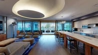 This elegantly designed single-floor space makes a striking first impression with its signature curved, recessed ceiling feature accented by warm ambient lighting — a defining architectural element that anchors the main lounge area. Wide-plank hardwood flooring flows throughout, unifying the various zones within the space.

The main lounge area offers generous seating arrangements, including plush sofas, tufted ottomans, and sculptural accent chairs in a mix of blue and warm neutral tones, set atop a bold patterned area rug. A wall-mounted flat-screen television and built-in media console make this zone ideal for both relaxed viewing and social gatherings. A secondary lounge area features swivel chairs in vibrant blue, arranged around a distinctive metallic accent table, lending an artistic, contemporary feel.

Floor-to-ceiling glass walls and sliding doors connect the interior to an outdoor terrace lined with lush greenery, flooding the space with natural light and providing open views of the surrounding cityscape. A dramatic sculptural chandelier hangs above a long marble-topped communal table with bar-height seating, perfect for collaborative or dining use.

The fully equipped kitchen is finished with light wood cabinetry, marble countertops and backsplash, an induction cooktop, double wall ovens, and integrated appliances — offering full culinary functionality within this upscale communal setting.