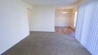 This single-floor residence offers a clean, open layout designed for comfortable everyday living. The main living area is finished with plush, neutral-toned carpeting that adds warmth underfoot, seamlessly transitioning into an adjoining space featuring rich hardwood-style flooring. The contrast between the two flooring types clearly delineates the living and dining areas while maintaining a cohesive aesthetic throughout.

The dining area is anchored by a classic chandelier that serves as a refined focal point, casting an inviting glow over the space. Natural light filters in through a window dressed with horizontal blinds, brightening the interior and enhancing the airy, open feel. The walls throughout are finished in a soft, neutral palette, providing a versatile backdrop that suits a wide range of furnishing styles.

The open-concept flow between the living and dining areas creates a spacious and functional layout, ideal for both everyday use and entertaining. The clean lines, understated finishes, and thoughtful details — from the decorative lighting to the dual flooring materials — give this home a move-in-ready appeal with a timeless, welcoming character.