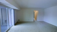This single-floor residence centers around a generously sized living area finished with wall-to-wall carpeting that adds warmth and comfort underfoot. The space is immediately distinguished by its expansive wall of sliding glass doors, which flood the interior with abundant natural light and provide direct access to a private balcony. The balcony features a white railing and offers pleasant views of snow-dusted evergreen trees and neighboring structures, creating a tranquil outdoor backdrop.

The living area is finished with smooth, light-colored walls and a clean white ceiling, contributing to a bright and airy atmosphere throughout. A ceiling light fixture provides overhead illumination, complementing the generous daylight entering through the large glazed doors. Sliding curtain panels frame the glass doors, allowing for adjustable light control and privacy.

A floor-level ventilation register is visible along the baseboard, indicating a built-in climate control system. An open doorway on the far wall leads to an adjacent hallway, connecting the main living space to the rest of the unit. The overall layout and neutral palette create a versatile, move-in-ready environment well-suited to a variety of furnishing arrangements.