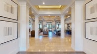 This expansive two-floor community amenity space offers a sophisticated blend of architectural elegance and functional design across a generously sized main floor with a secondary upper level. The grand entrance hallway sets the tone immediately, featuring tall white columns, polished travertine tile flooring, coffered ceilings with warm recessed lighting, and a stunning circular chandelier that anchors the open-concept layout. Large windows and glass doors throughout flood the interior with natural light while framing outdoor views.

Multiple living rooms provide inviting gathering spaces furnished with contemporary seating and curated artwork. The dining areas and kitchens are thoughtfully appointed, including a stylish bar nook with a chevron-patterned tile accent wall, dark cabinetry, and built-in beverage cooler. An intimate lounge area features bold, artistic wall coverings and gallery-style artwork, creating a refined retreat atmosphere.

The dedicated office space offers workstations alongside a separate conference-style meeting room with shiplap walls and ample natural light. The fitness center is expansive and well-equipped with cardio machines, free weights, strength equipment, ceiling fans, and mirrored walls. Outside, a resort-style pool with in-water lounge chairs, stone accent walls, and shaded patio seating areas completes this impressive amenity-rich space.