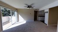 This single-floor space offers a practical and comfortable layout with a seamless flow between the main living area and kitchen. The living room is bathed in natural light streaming through large sliding glass doors, which open to an outdoor area and cast bright, airy light across the carpeted floor. A ceiling fan with a built-in light fixture is centrally mounted, providing both air circulation and overhead illumination. Neutral-toned walls throughout give the space a clean, understated aesthetic.

The kitchen is visible from the living area, featuring white upper cabinetry, dark countertops, and white appliances, including a dishwasher. The kitchen's darker flooring contrasts with the carpeted living space, creating a natural visual boundary between the two areas. A hallway leads further into the unit, connecting the main living spaces to additional rooms beyond. HVAC venting is integrated into the ceiling, ensuring efficient climate control throughout. The overall layout is open and functional, making efficient use of the available space while maintaining a bright and welcoming atmosphere.