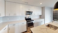 This single-floor residence offers a thoughtfully designed open-concept layout with two bedrooms and two and a half bathrooms. The living area is bright and spacious, finished with light wood-tone flooring and neutral gray walls. Large windows and a sliding glass door open to a private balcony, flooding the space with natural light and providing elevated views of the surrounding cityscape.

The kitchen is outfitted with white flat-panel cabinetry extending to the ceiling, stainless steel appliances including a French door refrigerator, electric range, and microwave, along with a central island featuring a quartz countertop with veining detail and a stainless steel sink. Pendant lighting accents the island, adding a contemporary touch.

Both full bathrooms feature light wood-look vinyl flooring, white vanity cabinetry, and dark stone-toned countertops. One includes a shower-tub combo with sliding glass doors, while another is accessed through a frosted glass door for added privacy. A half bath provides additional convenience.

The bedrooms are carpeted and generously sized, with large windows framing city views. The primary bedroom includes a sliding door to the balcony and an expansive walk-in closet outfitted with adjustable shelving and hanging rods.