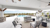 Located in O'Fallon, Missouri, this single-level community amenity space centers around a generously sized rectangular swimming pool enclosed by a black wrought-iron fence. The pool deck is finished in light-toned concrete and lined with numerous lounge chairs, offering ample space for sunbathing and relaxation with open views of the surrounding neighborhood and sky.

Adjacent to the pool is an expansive covered pavilion that serves as the heart of the outdoor living area. This covered space features a tongue-and-groove white ceiling with multiple ceiling fans to ensure comfortable airflow. The pavilion is anchored by a stone-clad fireplace with a rustic wood mantel and a wall-mounted television above, creating an inviting gathering spot for all seasons. Plush outdoor sofas and lounge chairs are arranged thoughtfully throughout, complemented by area rugs and side tables.

A separate outdoor kitchen area with a built-in grill adds functional appeal for outdoor entertaining. The pavilion transitions seamlessly to an al fresco dining zone, complete with bar-height seating. The overall design aesthetic is clean and contemporary, with a neutral gray and white palette that lends a polished, cohesive look. The combination of shaded comfort and open-air recreation makes this a well-appointed community destination.