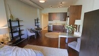 This well-appointed open-concept studio showcases a thoughtfully designed layout that maximizes every square foot across a single floor. Rich, dark hardwood flooring flows throughout the entire space, lending warmth and continuity to the interior. The kitchen is outfitted with deep espresso-toned cabinetry, stone tile backsplash, granite countertops, and stainless steel appliances including a dishwasher and refrigerator, creating a sleek and functional cooking area. Track lighting above the kitchen ensures excellent task illumination, while a ceiling-mounted light fixture adds to the ambient glow of the space. The open layout transitions naturally into the dining area, where a compact round table with upholstered chairs offers an intimate setting. A modern rectangular dining or work table with a metallic surface provides additional versatility. Decorative touches such as a tiered display shelf, fresh white floral arrangements, and layered area rugs — including a plush shag rug in the living zone — add texture and personality throughout. Exposed ductwork along the ceiling contributes an industrial design element that complements the contemporary finishes. The cohesive, open arrangement of living, dining, and kitchen areas makes this a highly functional and visually appealing single-level space.