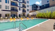 This expansive outdoor courtyard amenity space is thoughtfully designed to offer a comprehensive resort-style experience. At its centerpiece sits a large, sparkling swimming pool surrounded by rows of yellow and green lounge chairs, providing ample space for sunbathing and relaxation beneath open skies. The pool deck flows seamlessly into a series of distinct outdoor zones, each serving a unique purpose.

A covered pavilion area features a warm wood-paneled ceiling supported by sleek steel pergola framing, creating a partially shaded retreat with comfortable modern seating. Large floor-to-ceiling glass folding doors connect the covered patio directly to an interior lounge, blurring the boundary between indoor and outdoor living. An adjacent turf lawn area is equipped for lawn games and casual gatherings, with a wall-mounted outdoor television for entertainment.

Throughout the courtyard, carefully curated landscaping including ornamental grasses, flowering shrubs, and bamboo plantings adds natural texture and greenery. A quieter seating alcove with modular lounge furniture offers a more intimate setting for conversation. The surrounding multi-story building facade, clad in white horizontal siding with bold navy blue accents and private balconies, frames the entire courtyard with a clean, contemporary aesthetic that maximizes natural light throughout the day.