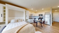 This thoughtfully designed studio unit offers a single-floor, open-concept layout that maximizes both functionality and comfort. The main living area centers around a built-in Murphy bed flanked by white cabinetry and shelving, allowing the space to transition seamlessly between a living and sleeping area. Light wood-style flooring runs throughout, lending warmth and continuity to the entire unit.

The kitchen is a standout feature, equipped with stainless steel appliances including a refrigerator, gas range, microwave, and dishwasher. White shaker-style cabinetry, a subway tile backsplash, dark countertops, and a wine rack accent above the upper cabinets give the kitchen a clean, polished character. A compact dining area with a round table sits adjacent to the kitchen, ideal for everyday meals.

A sliding glass door opens to a private balcony, filling the living area with natural light and offering open outdoor views. A wall-mounted television adds to the unit's functional design.

The bathroom is finished with a rich wood-toned vanity, dual undermount sinks, a backlit mirror, patterned tile flooring, and a frameless glass walk-in shower, creating a spa-like atmosphere. A smart thermostat is also featured in the hallway, adding a modern, convenient touch to daily living.