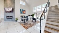 Located at 4020 S 130th East Ave in Tulsa, Oklahoma, this single-floor space makes a striking impression with its dramatic vaulted ceilings that amplify the sense of openness and volume throughout. Large clerestory and floor-to-ceiling windows flood the interior with natural light, creating a bright, airy atmosphere against a crisp white palette of walls and large-format tile flooring.

The living area is thoughtfully designed, anchored by a bold navy blue channeled sofa, sculptural coffee tables, and a pair of cream accent chairs arranged atop a richly textured area rug. A stone-clad accent wall houses a sleek linear fireplace with a wall-mounted television above, adding both warmth and a focal point to the lounge area. Curated wall art and decorative 3D panels contribute to the space's contemporary aesthetic.

Adjacent to the lounge, a dining and gathering area features multiple seating arrangements with modern chairs around round tables, alongside a service counter or kitchenette station. Lush indoor plants punctuate the space throughout, bringing life and color to the clean, modern design. A staircase with cable railings leads to an upper level, further enhancing the architectural interest. The overall ambiance is sophisticated, welcoming, and well-suited for both relaxation and social gatherings.