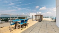 Located at 1 Harbor Point Road in Stamford, Connecticut, this rooftop terrace offers a well-appointed outdoor retreat set high above the surrounding cityscape. The space is finished with large-format stone pavers throughout, providing a clean, modern aesthetic that extends across the entire level.

The terrace is divided into distinct functional zones. A dedicated dining area features a high-top table with seating for several guests, positioned along the glass-panel perimeter railing to maximize unobstructed views of Stamford and beyond. A slatted wood-and-metal pergola structure nearby provides a shaded seating option, adding architectural interest to the open-air layout.

A separate lounge zone incorporates artificial turf, offering a softer surface for relaxation. Light blue chaise lounges are arranged here for sunbathing, backed by a contemporary panel wall and a manicured hedge accent. This section is ideal for casual outdoor recreation away from the dining area.

Throughout the terrace, glass-panel railings maintain safety while preserving sweeping panoramic views that stretch across the Stamford skyline, surrounding greenery, and toward Long Island Sound. The open sky above and abundant natural light make this rooftop an inviting, versatile outdoor space suited for both social gatherings and quiet relaxation.