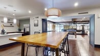Located at 110 S Hoover Blvd in Tampa, FL, this thoughtfully designed single-floor amenity space blends modern aesthetics with functional communal living. Large-format tile flooring runs throughout, anchoring the open layout and connecting each distinct zone seamlessly.

The kitchen is a standout feature, outfitted with rich dark wood cabinetry, white quartz countertops, a central island with pendant lighting, stainless steel appliances including a French door refrigerator and dishwasher, and a deep undermount sink. Open wood shelving against a geometric patterned accent wall adds both storage and visual interest.

A spacious dining area adjoins the kitchen, centered around a solid wood communal table beneath a woven rattan pendant light, offering ample seating for group gatherings. The lounge area provides a relaxed retreat, furnished with an olive green velvet sofa, a round coffee table, and layered rugs set against a bold geometric wallpapered accent wall complemented by botanical artwork.

Beyond the main living areas, a glass-partitioned recreation room houses a ping-pong table and additional game space, with natural light filtering in through large windows. Recessed lighting throughout ensures a bright, welcoming atmosphere across the entire space.