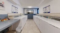 Located at 1202 Harwood St in Vancouver's West End, this shared laundry facility is a well-organized, single-floor amenity space designed for efficient, communal use. The room is laid out with two parallel rows of full-sized, commercial-grade top-load washing machines and dryers lining both sides, providing ample capacity for residents. Light-toned large-format tile flooring runs throughout, lending the space a clean and practical aesthetic. A blue-grey ceramic tile wainscoting accents the lower portion of the walls, adding a touch of visual character. A built-in folding counter runs along one side of the room, offering a convenient surface for sorting and folding laundry. At the far end of the room, a large mirror is mounted above the tile wainscoting, enhancing the sense of depth in the space. Overhead flush-mount lighting provides consistent illumination throughout. A wall-mounted payment terminal near the entrance allows for easy, cashless access to the machines. Informational posted notices and a corkboard keep residents informed of facility guidelines. A seating area with a patterned chair near the entry door provides a comfortable spot to wait. The space is accessed through a solid grey door and includes a fire extinguisher, reflecting attention to safety standards.