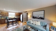 This one-bedroom, one-bathroom unit at The District in University City, Missouri offers a well-designed, single-floor layout that makes efficient use of its space. The open-concept living and dining area is anchored by warm hardwood-style flooring, neutral taupe walls, and a textured ceiling, creating an inviting and cohesive atmosphere. A large window floods the living room with natural light, complementing the relaxed seating arrangement and media console setup.

The kitchen flows seamlessly from the living area and features rich, dark wood cabinetry, granite countertops, stainless steel appliances including a full-size refrigerator and dishwasher, and a double stainless steel sink beneath a window with outdoor views. Open wooden shelving adds both functionality and a warm, rustic accent to the space.

A hallway with hardwood-style floors and recessed lighting connects the main living area to the private bedroom and bathroom. The bedroom offers plush carpeting, a bold accent wall in deep blue-gray, and a window with views of the surrounding greenery, providing a calm and restful retreat.

The bathroom is clean and functional, featuring white tile walls, a tub-shower combination, a pedestal sink with brushed nickel fixtures, and a framed mirror. The overall design throughout the unit is cohesive, modern, and comfortable, making it a practical and stylish home in a convenient University City location.
