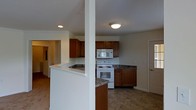 This single-floor residential space is designed with functionality and comfort in mind. The kitchen serves as a central highlight, featuring rich warm-toned wood cabinetry that extends from base to upper wall units, providing generous storage throughout. Dark granite-style countertops offer a practical and attractive contrast to the lighter cabinetry and walls. The kitchen is equipped with a white electric range, an over-the-range stainless steel microwave, and ample counter space for meal preparation. A structural support column adds architectural character to the open layout, while a peninsula-style counter section creates a natural division between the kitchen and the adjoining living areas. The flooring throughout the kitchen and connecting spaces features durable tile in a neutral, earthy tone, contributing to a cohesive and easy-to-maintain aesthetic. Recessed and flush-mount ceiling lighting provides consistent illumination throughout the space. Wide doorways connect the kitchen to additional rooms, hinting at a thoughtfully laid-out floor plan. The overall ambiance is clean, practical, and welcoming, making this a well-appointed space suited for everyday living.