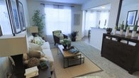 This single-floor space features a thoughtfully designed living area centered around a natural aesthetic. The living room is anchored by a plush white sofa adorned with botanical-patterned cushions, complemented by dark wood armchairs that add contrast and warmth. A wooden coffee table with a metal frame serves as the focal point, styled with decorative plants and books atop a woven area rug that grounds the seating arrangement. Large windows dressed in sheer white curtains allow generous natural light to fill the room, creating an airy, welcoming atmosphere. Tall indoor plants, including a striking bamboo arrangement, reinforce the nature-forward design throughout. The flooring transitions from carpet in the living area to hard surface flooring toward the adjacent space. A half-wall partition subtly separates the living room from what appears to be a kitchen or dining area, maintaining an open, connected feel while defining each zone. The overall ambiance is calm and cohesive, with a consistent green-and-neutral color palette that ties the botanical décor together seamlessly.