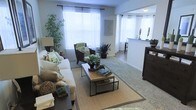 This single-floor space features a thoughtfully designed living area centered around a natural aesthetic. The living room is anchored by a plush white sofa adorned with botanical-patterned cushions, complemented by dark wood armchairs that add contrast and warmth. A wooden coffee table with a metal frame serves as the focal point, styled with decorative plants and books atop a woven area rug that grounds the seating arrangement. Large windows dressed in sheer white curtains allow generous natural light to fill the room, creating an airy, welcoming atmosphere. Tall indoor plants, including a striking bamboo arrangement, reinforce the nature-forward design throughout. The flooring transitions from carpet in the living area to hard surface flooring toward the adjacent space. A half-wall partition subtly separates the living room from what appears to be a kitchen or dining area, maintaining an open, connected feel while defining each zone. The overall ambiance is calm and cohesive, with a consistent green-and-neutral color palette that ties the botanical décor together seamlessly.
